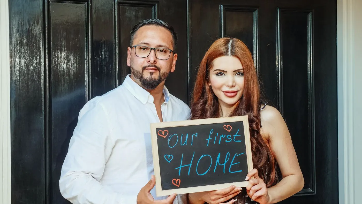A smiling couple at the door of their new home holding a sign that reads Our first home